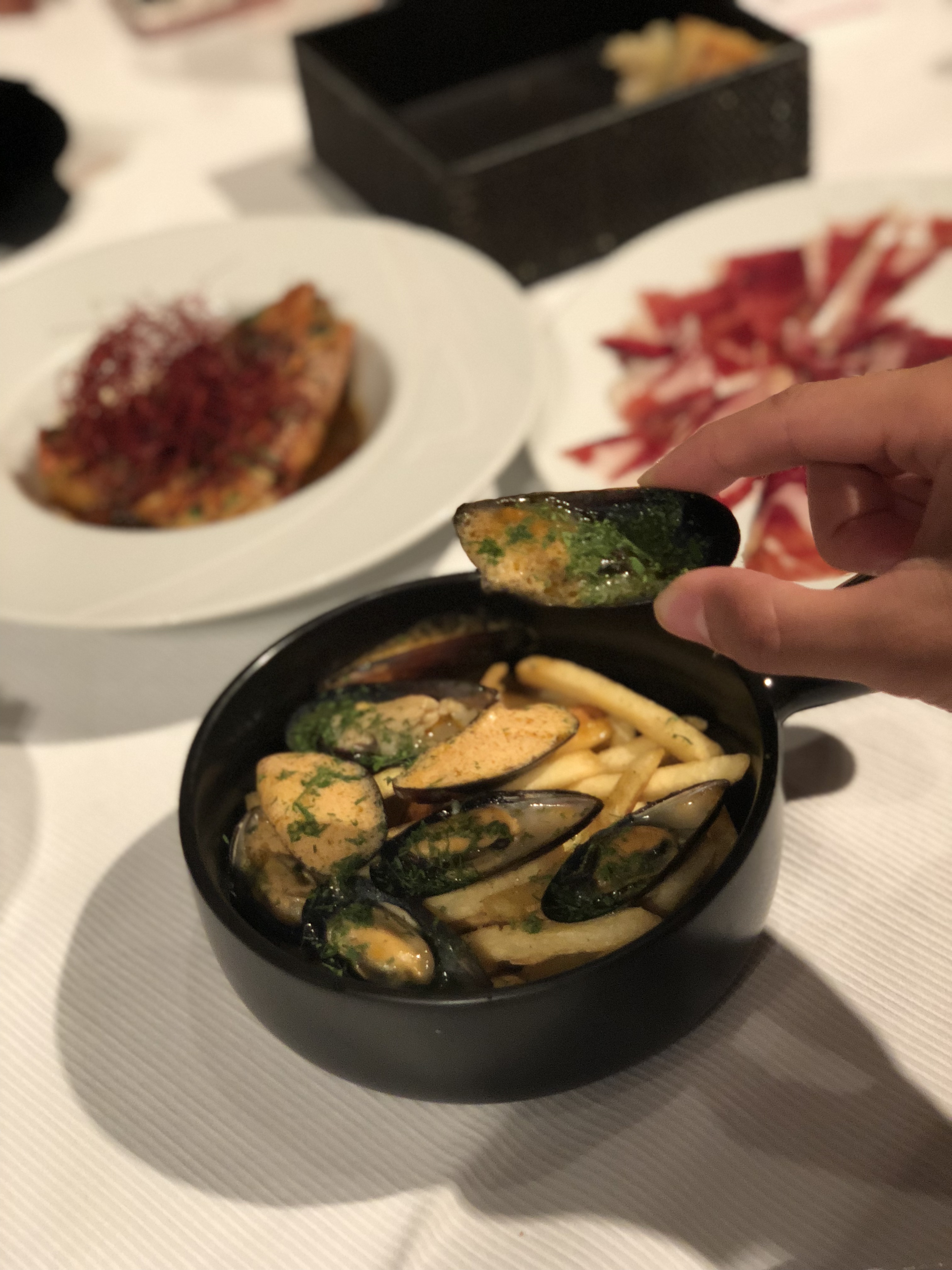 french fries with mussels, hand holding one mussel. Dishes in the background